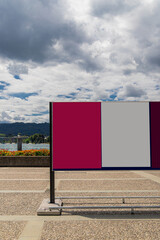 Mock-up of blank red and gray billboard on lakeside promenade with mountains and cloudy sky.