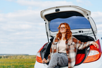 young beautiful redhead caucasian woman sits on edge of car trunk, white passenger car standing on...