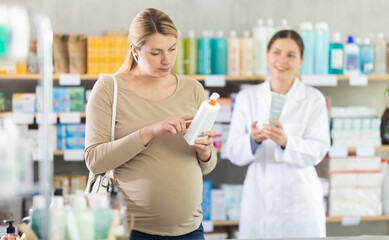 Pregnant woman choosing cream with help of young female pharmacist in pharmacy