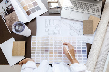 Top view of interior designer or architect in casual wear working on a blueprint for new project, standing in her office on a daytime. Laptop, color samples, coffee cup, tools