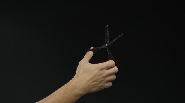 Man holding black scissors on isolated dark background, highlighting male hand and tool in a minimalist setting with a focus on craftsmanship and detail contrast.