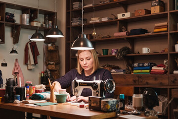 Caucasian young adult woman working at sewing machine in workshop, focusing on fabric project with shelves of textiles and supplies in background, hands engaged in precise stitching