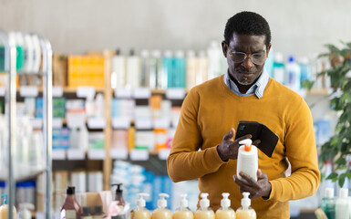 Focused middle aged african american man in casual ochre sweater and glasses scanning qr code on body lotion package(bottle, tube) with phone, browsing product information online in pharmacy..
