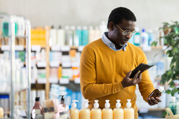 Interested middle-aged black man in mustard yellow casual sweater pharmacy examining brown cough syrup bottle while using smartphone with interest to scan label to verify ingredients or dosage online