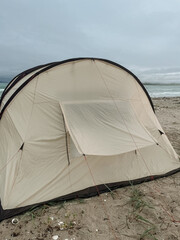 A beige tent set up on a sandy beach near the ocean. The sky is overcast, and gentle waves can be seen in the background.