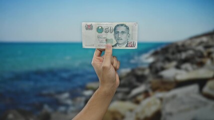 Man holding singapore dollar banknote against a picturesque beach, highlighting travel and financial freedom in a serene seaside environment.