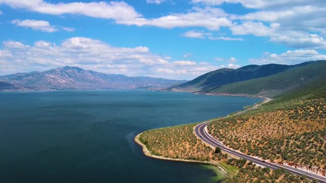An aerial view captures the breathtaking beauty of Bafa Lake in Turkey, showcasing a winding coastal road alongside its tranquil, deep blue waters. Majestic mountains rise in the background under a cl