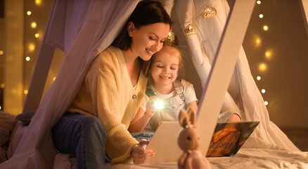 Cute little girl with flashlight and her mother reading story in hut at night