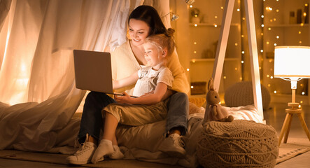 Cute little girl with her mother watching cartoons on laptop in bedroom at night © Pixel-Shot