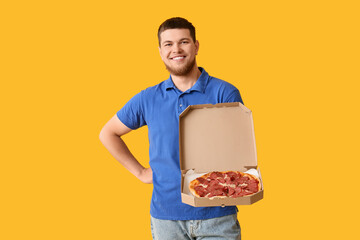 Happy young man holding cardboard box with tasty pepperoni pizza on yellow background