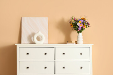 Vase with wild flowers, picture and decor on chest of drawers near beige wall in room