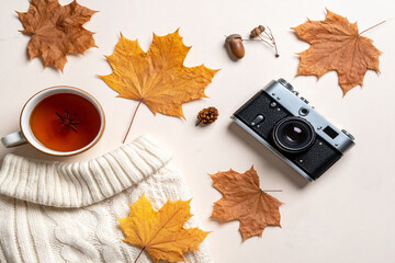 Cozy autumn flat lay with a vintage camera, tea cup, maple leaves, and a warm knitted scarf on a soft beige background.