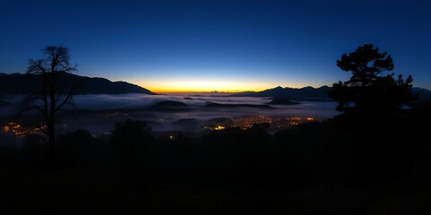 Panoramic view of a town nestled in a valley covered by fog at twilight with silhouetted trees in the foreground