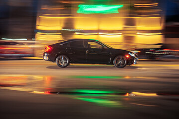 Black sedan car driving along illuminated wet street oat night. Long exposure. Bright colorful neon city lights blurred in background.
