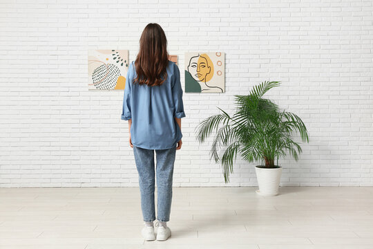 Young woman looking at paintings on white brick wall in art gallery, back view