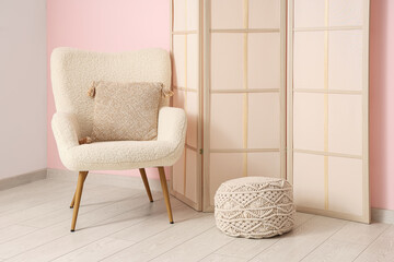 Soft armchair with pouf and folding screen near pink wall in room