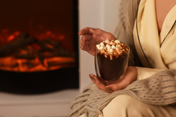 Beautiful young woman with cup of hot chocolate and marshmallows near fireplace in evening at home, closeup