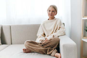 A young girl sits cross legged on a light sofa, wearing an oversized cream sweater and brown corduroy pants, relaxed in a sunlit, cozy living space.