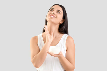 Young woman applying facial cream on light background