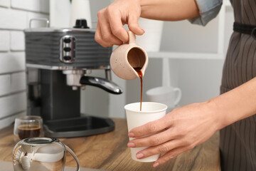 Young female barista pouring coffee from jug into disposable cup in coffee shop