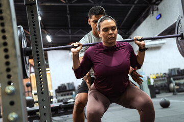 Latin american woman performing barbell squats at gym, personal trainer assisting with form and technique. Fitness and workout concept