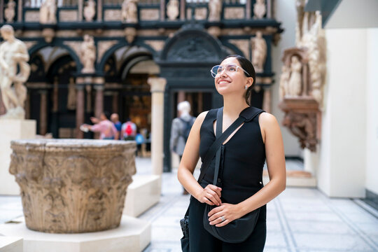 Young Latina woman with a bright smile looks up admiring sculptures inside the Victoria and Albert Museum in London, England. Wearing a modern outfit, she expresses curiosity joy in a artistic indoor