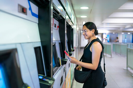 Young smiling Latina woman at a London Underground station in England pays for metro access using a digital card on her smartphone. She wears black clothes, a black bag while commuting - Powered by Adobe