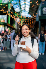 Young happy Latina woman enjoys trendy strawberries at Borough Market in London, England, she smiles joyfully in this iconic food destination filled with flavor, color, and a lively crowd
