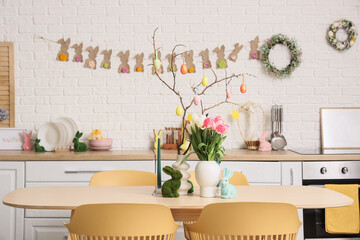 Interior of festive kitchen with tulip flowers, rabbits and Easter painted eggs on tree branches at dining table