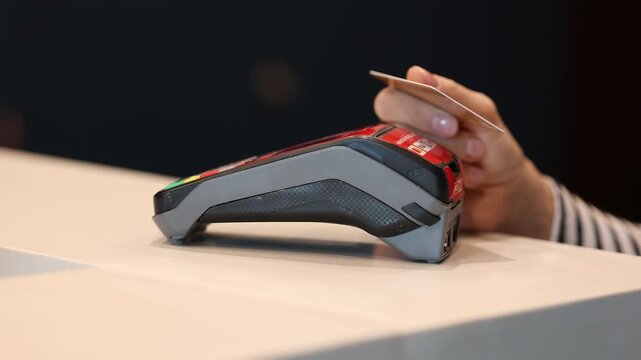 Close-up of female hand holding bank card near POS terminal for contactless payment. Concept of cashless transaction, shopping, and modern finance.