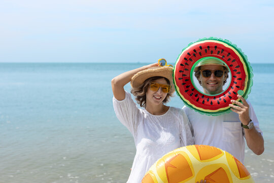 Smiling couple enjoying summer vacation at the beach, holding colorful fruit inflatable rings, wearing sunglasses and straw hats on a sunny tropical day.