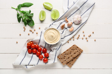 Bowl of tasty hummus with ingredients, crackers and napkin on white wooden background