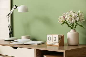 Cube calendar with date 3 February, book, flowers and lamp on wooden table near color wall