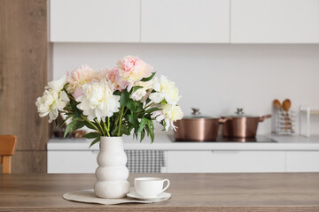 Vase with bouquet of beautiful peonies and cup of tea on table in kitchen, closeup