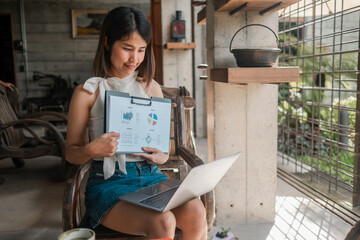 A young woman reviews business charts on a laptop and clipboard in a stylish industrial workspace.