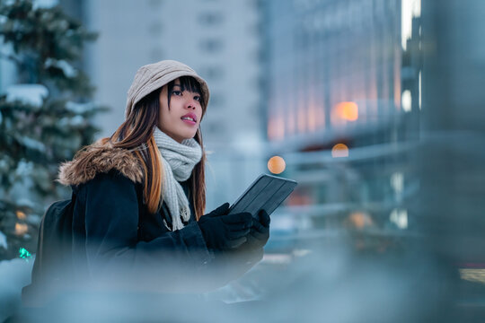 Asian woman remote working freelance on digital tablet while waiting for bus in winter night. Woman traveler enjoy urban lifestyle travel in the city with using wireless technology and gadget device.