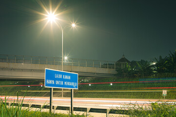 A blue sign stands illuminated by streetlights at night, with blurred car light trails streaking across the road beneath a bridge.