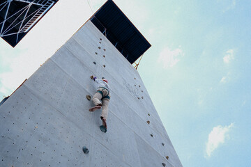 A person is climbing a tall, artificial rock climbing wall outdoors against a bright, partly cloudy sky.
