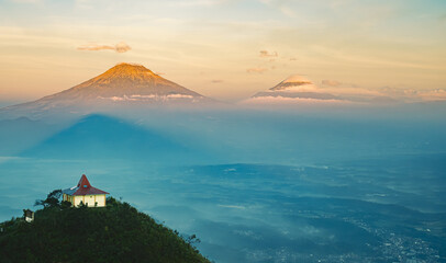 A small building sits atop a hill overlooking a valley with distant mountains shrouded in mist during a warm, golden sunrise.