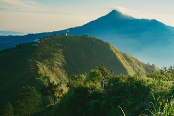 Scenic view of a lush green mountain range with a taller, hazy peak in the background under a clear blue sky.