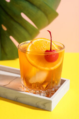 Glass of tasty Old Fashioned cocktail with cherry and monstera leaf on yellow table against beige background