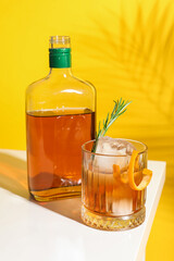 Glass of tasty Old Fashioned cocktail with rosemary on white table against yellow background