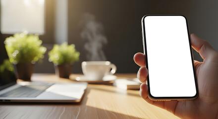 A person's hand holding a smartphone with a blank white screen, perfect for mockups, over a wooden desk with a laptop and coffee.