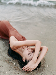 A young woman lies on a sunlit beach, body relaxed and arched along the sand, hair spread, water lapping nearby, creating a serene, intimate moment.
