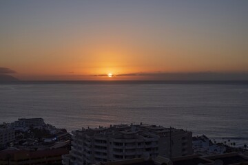 Sunset over ocean and Tenerife coastline. Scenic seascape with city buildings, calm Atlantic waters and orange sky. Canary Islands travel destination and evening nature background.