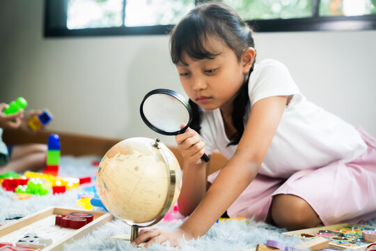 A young girl is looking at a globe with a magnifying glass. She is sitting on the floor surrounded by toys