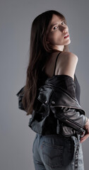 Fashion model with long brown hair wearing black leather jacket and jeans posing in studio, looking over shoulder