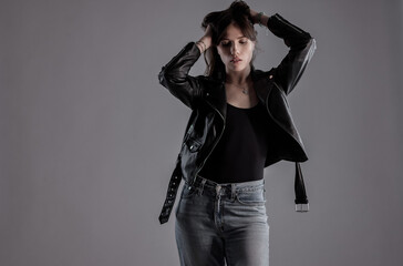 Studio portrait of a fashion model posing with hands on her hair, wearing a black leather jacket and jeans