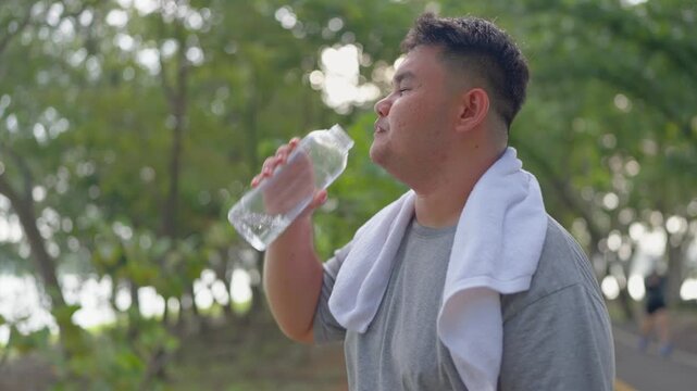 Asian plus-size man drinking water while exercises outdoors in garden.