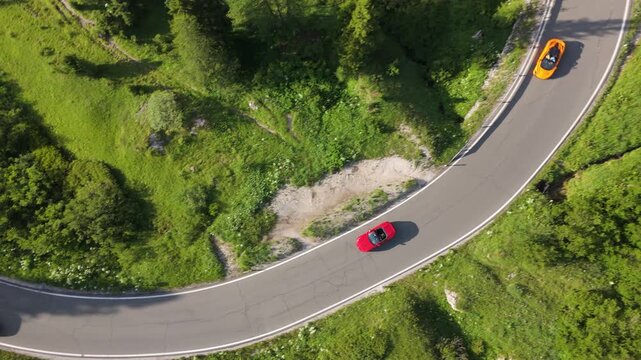 Drone aerial shot of luxury sports cars entering a sharp hairpin turn on a mountain serpentine road. Scenic alpine landscape. Category: Transportation